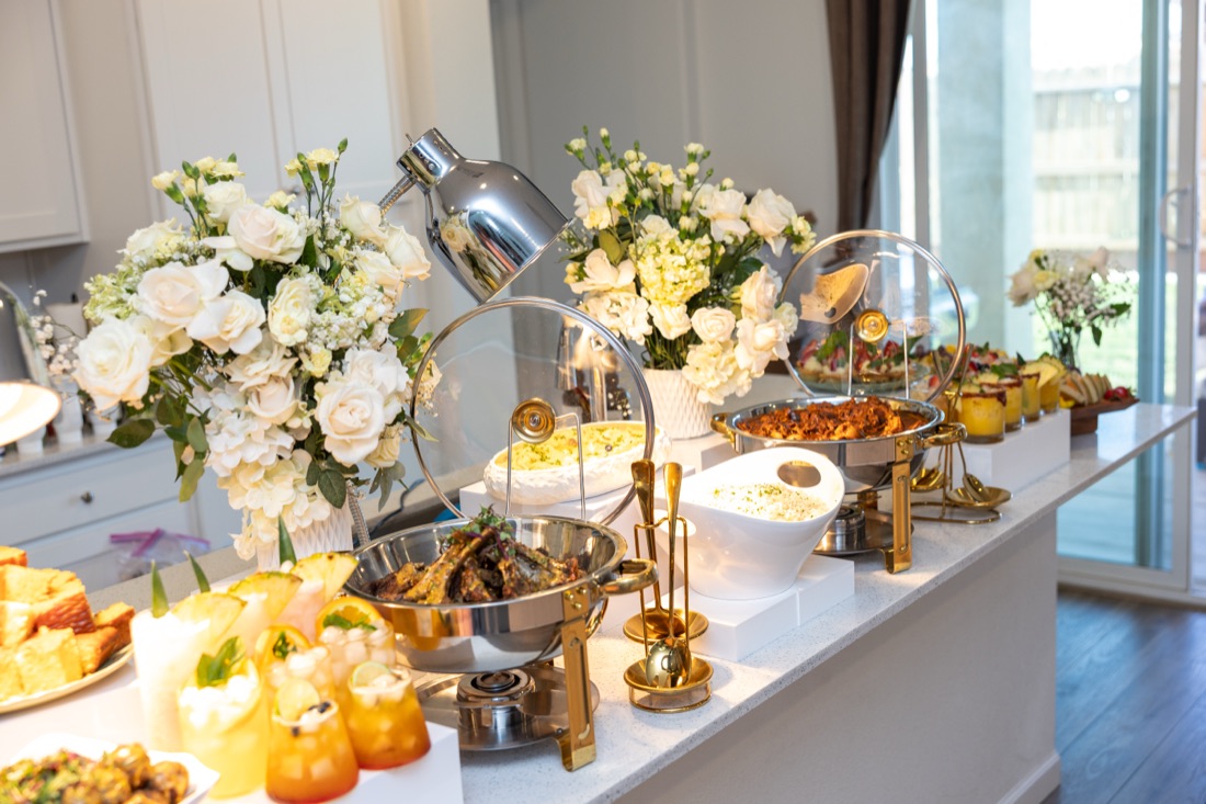 Buffet line of silver-and-gold chafing dishes with white rose arrangements, dessert display, and heat lamp on a bright kitchen island