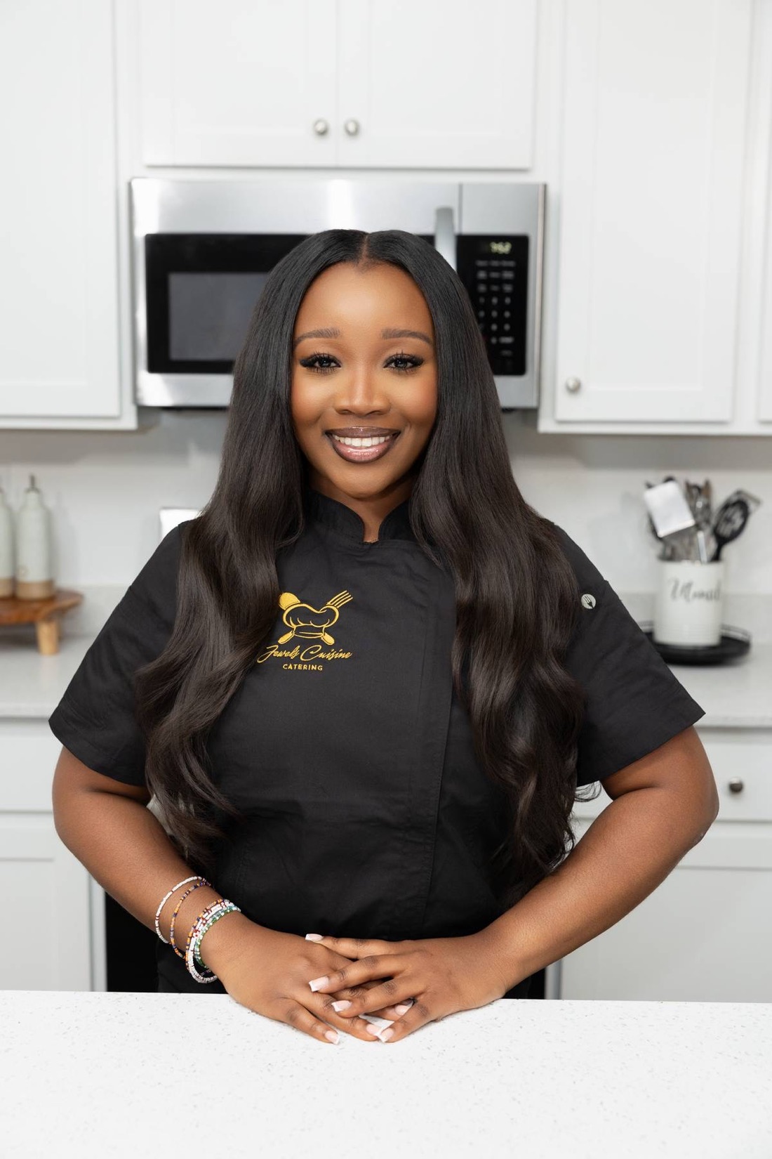 Professional portrait of Chef Jewel Brown in a black Jewel's Cuisine chef coat smiling in a bright white kitchen