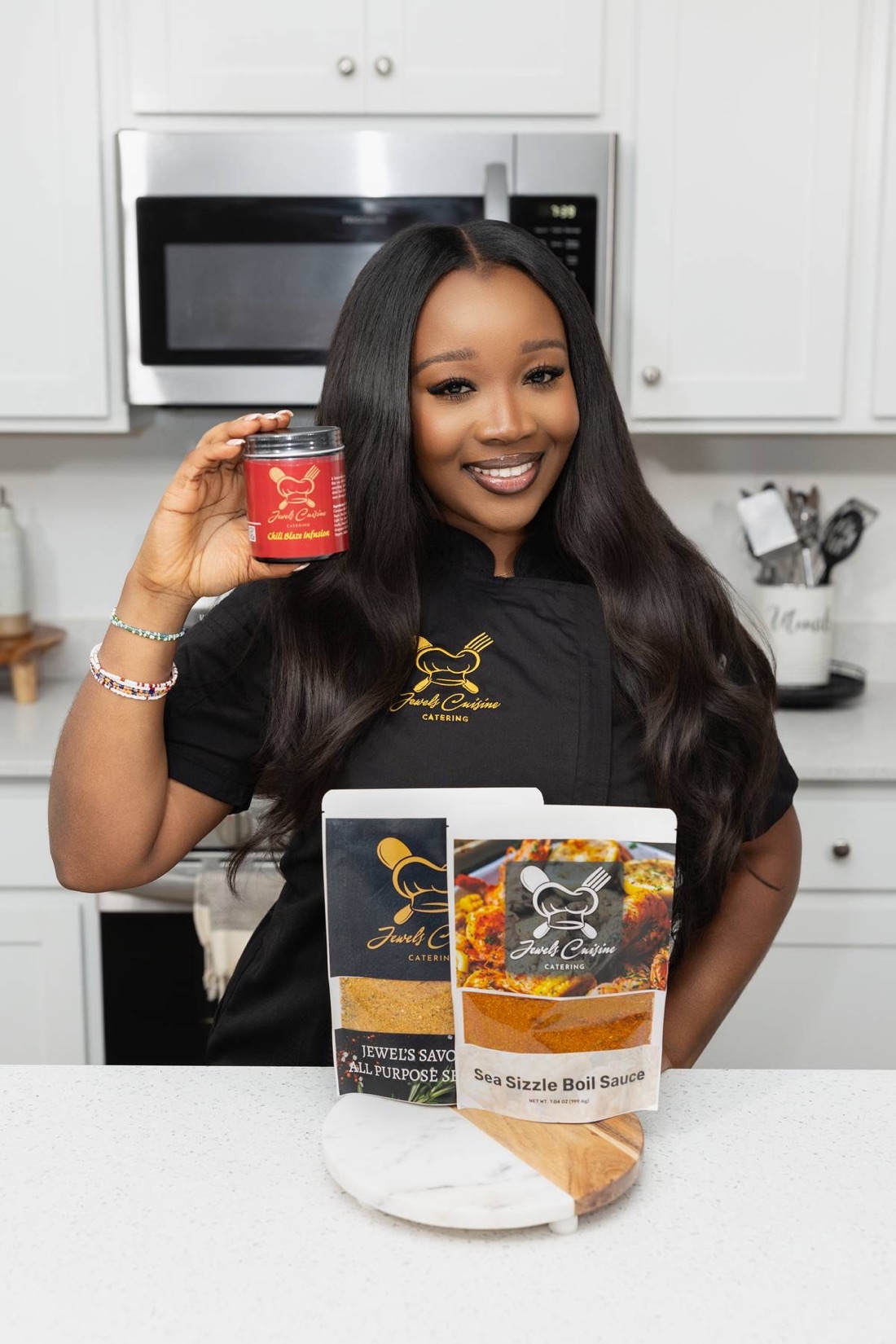 Chef Jewel Brown in a black chef coat smiles holding a Chili Blaze Infusion jar with Savory Mix and Sea Sizzle Boil Sauce pouches displayed on the counter