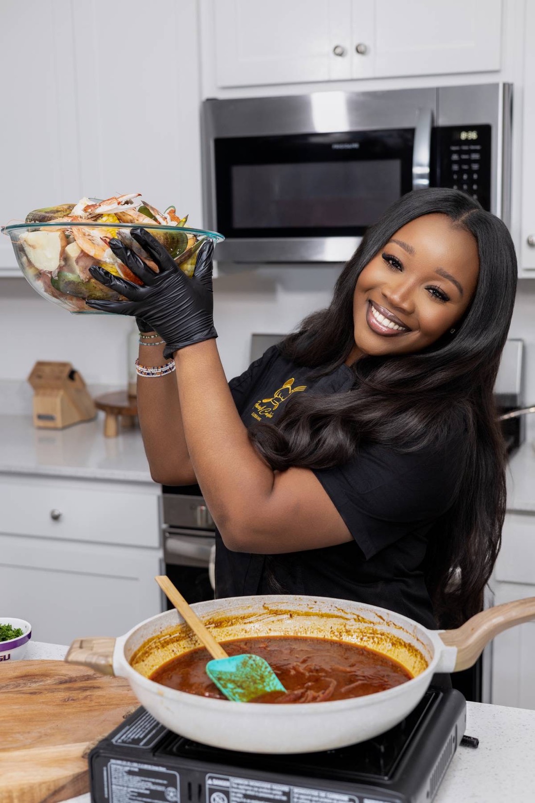 Chef Jewel Brown lifts a bowl of seafood boil ingredients over a simmering pan of red boil sauce, smiling in a bright kitchen