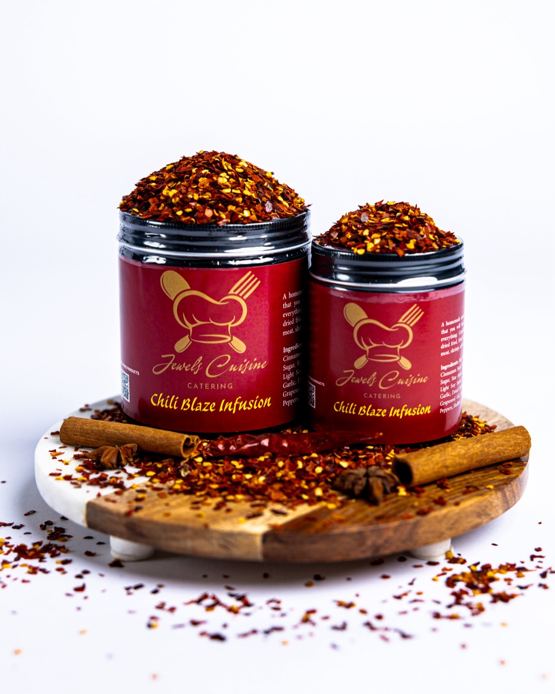 Two red jars of Jewel's Cuisine Chili Blaze Infusion seasoning overflowing with chili flakes on a marble-and-wood board with cinnamon sticks