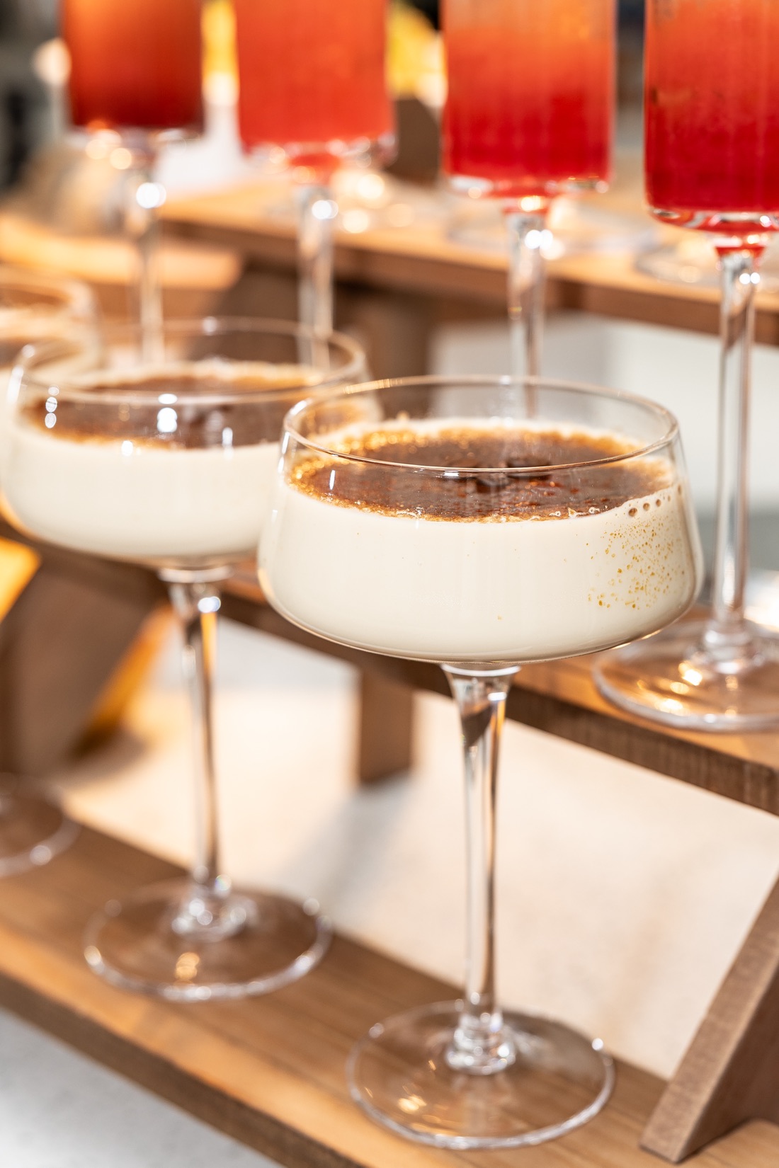 Coupe glasses of creamy crème brûlée-style dessert with caramelized sugar tops on a tiered wood stand, red drinks behind