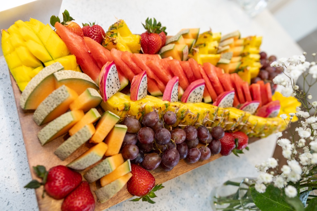 Overhead fruit board with mango, watermelon, cantaloupe, pineapple, dragon fruit, grapes, and strawberries with baby's breath florals