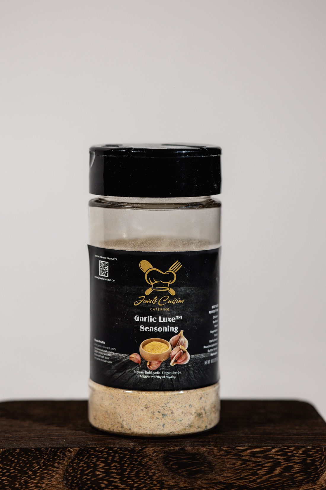 Studio product shot of Jewel's Cuisine Garlic Luxe Seasoning jar with black and gold label on a dark wood board