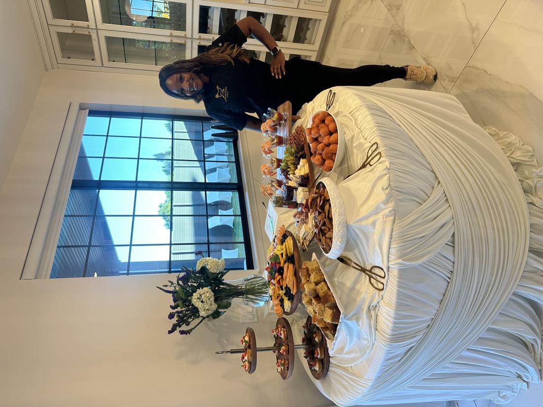 Chef Jewel Brown standing proudly beside a fully styled grazing table with shrimp cocktail, charcuterie, and desserts