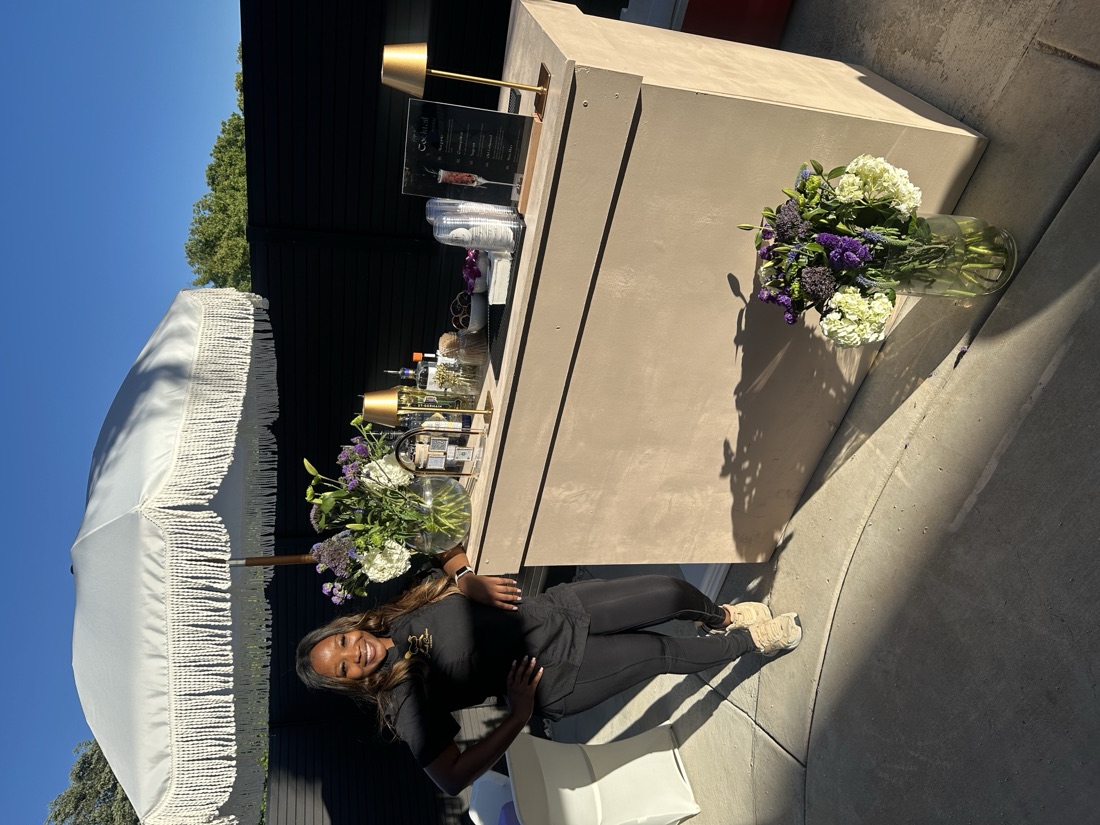 Chef Jewel Brown in black smiling beside her cream mobile bar under a white fringe umbrella at an outdoor event