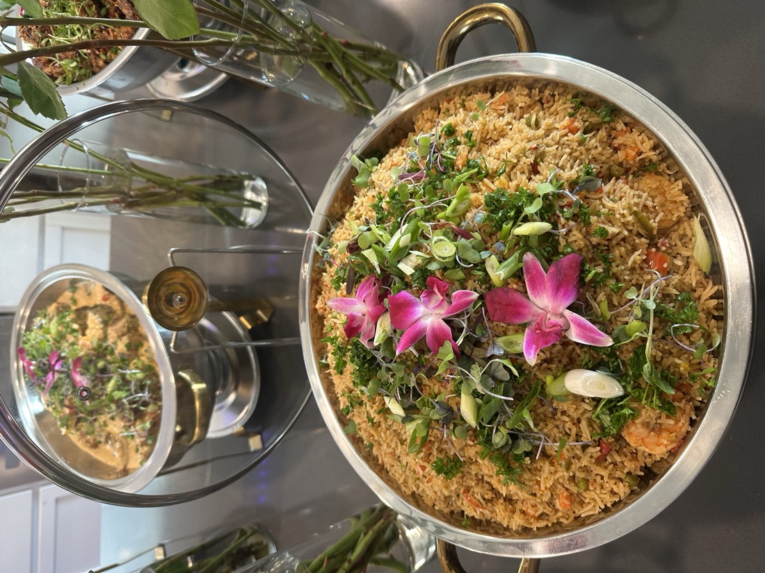 Chafing bowl of Liberian-style jollof rice with shrimp, crowned with microgreens, scallions and pink orchids