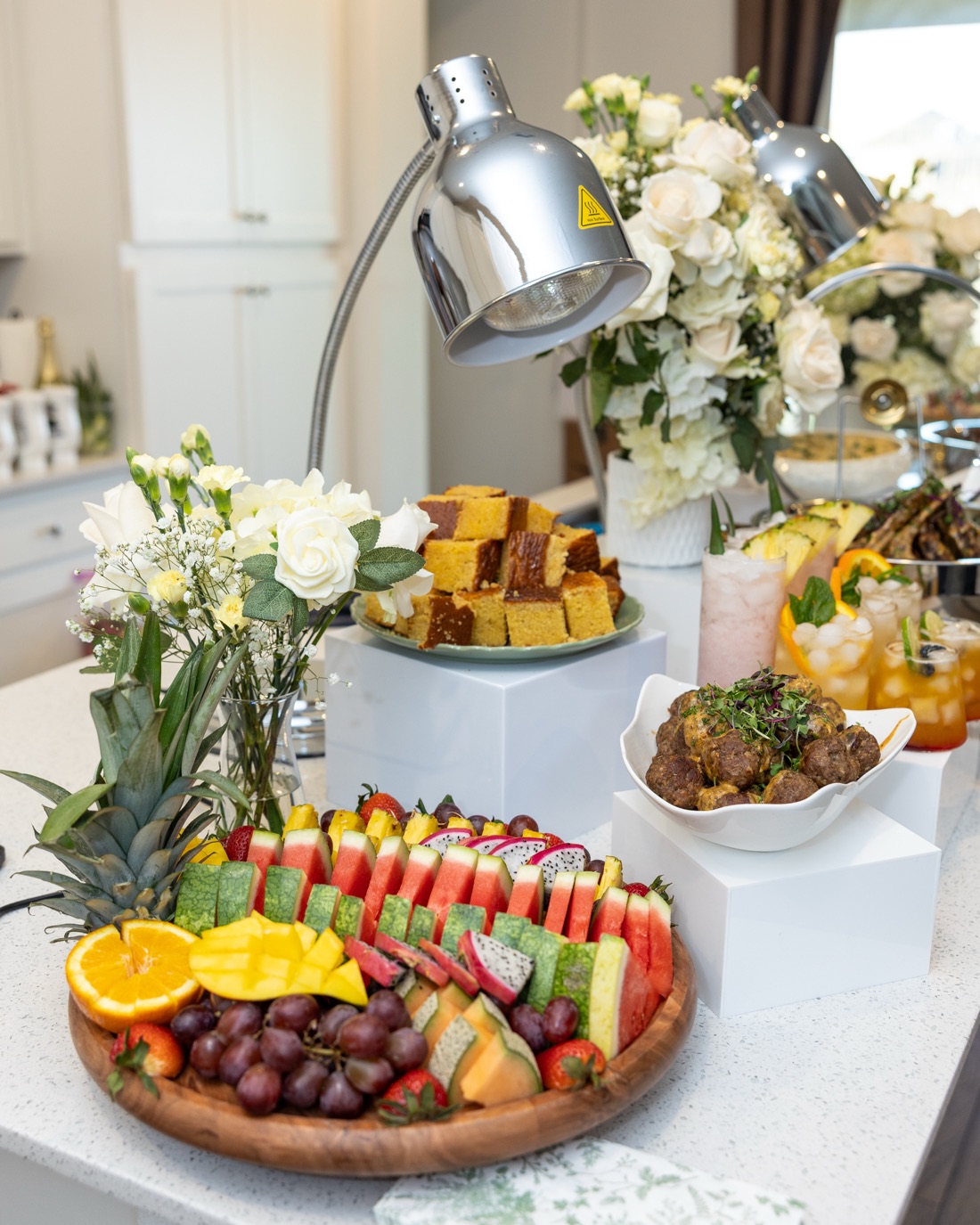 Full luxury brunch buffet with chrome heat lamps, stacked cornbread, glazed meatballs, tropical fruit platter, and white rose arrangements