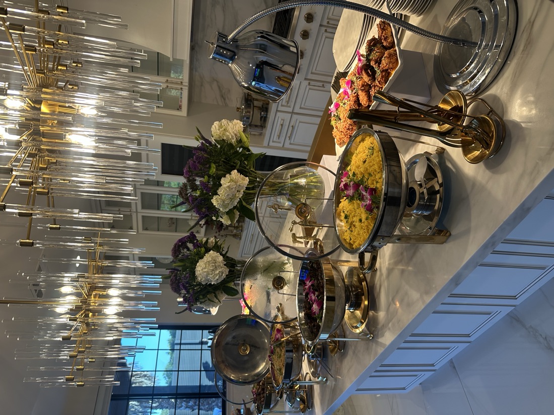 Long marble kitchen island buffet with polished chafing dishes of rice and entrees beneath glass-and-gold chandeliers
