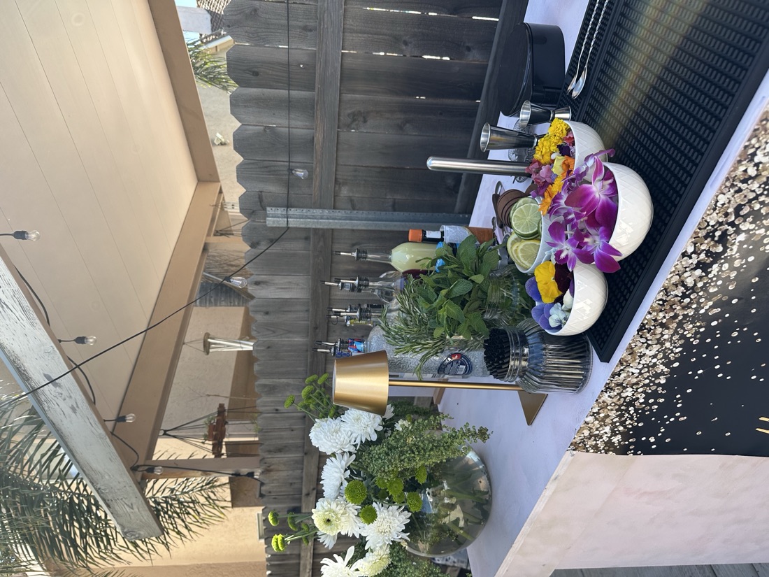 Outdoor mobile bar garnish station with bowls of edible flowers, citrus wheels, fresh mint, gold cordless lamp and white floral arrangement