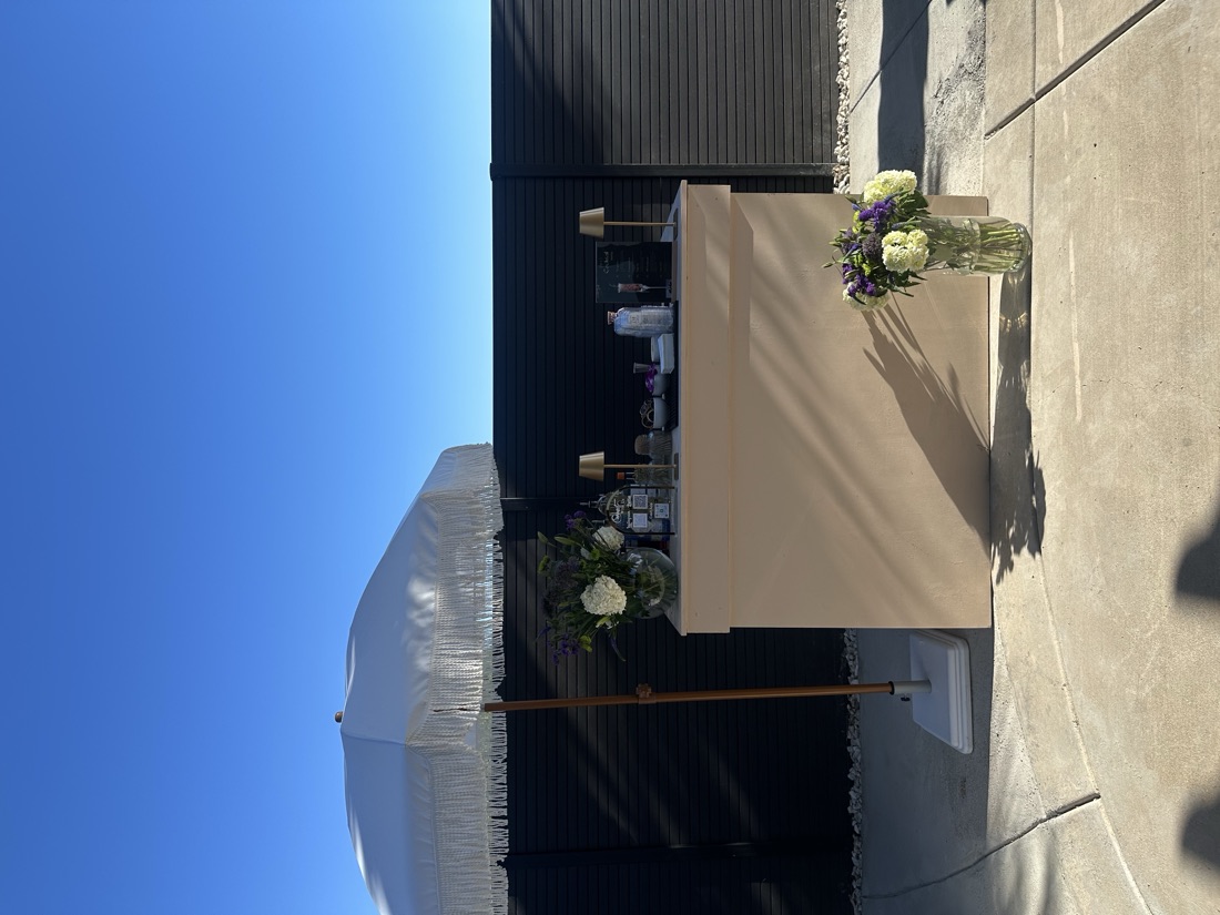 Cream mobile bar with a white fringe umbrella and fresh florals set against a black fence under a clear blue sky