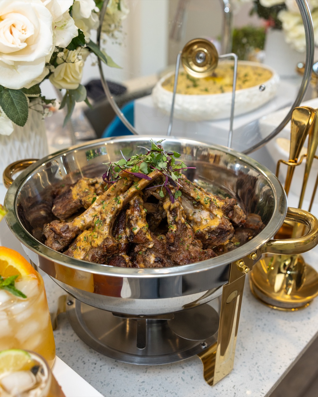 Herb-sauced lamb chops topped with microgreens in a silver-and-gold chafing dish beside white roses on a luxury buffet