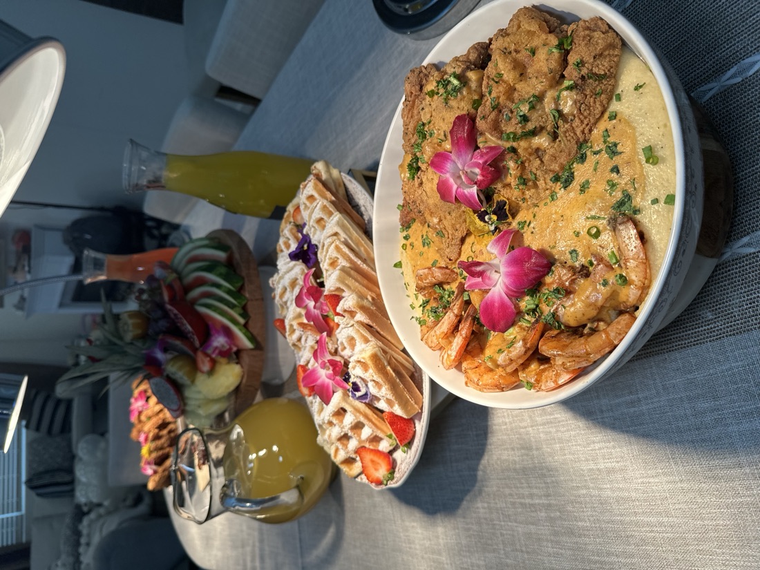 Hero bowl of creamy shrimp and grits with fried chicken, garnished with orchids, in front of a full brunch spread with waffles and fruit