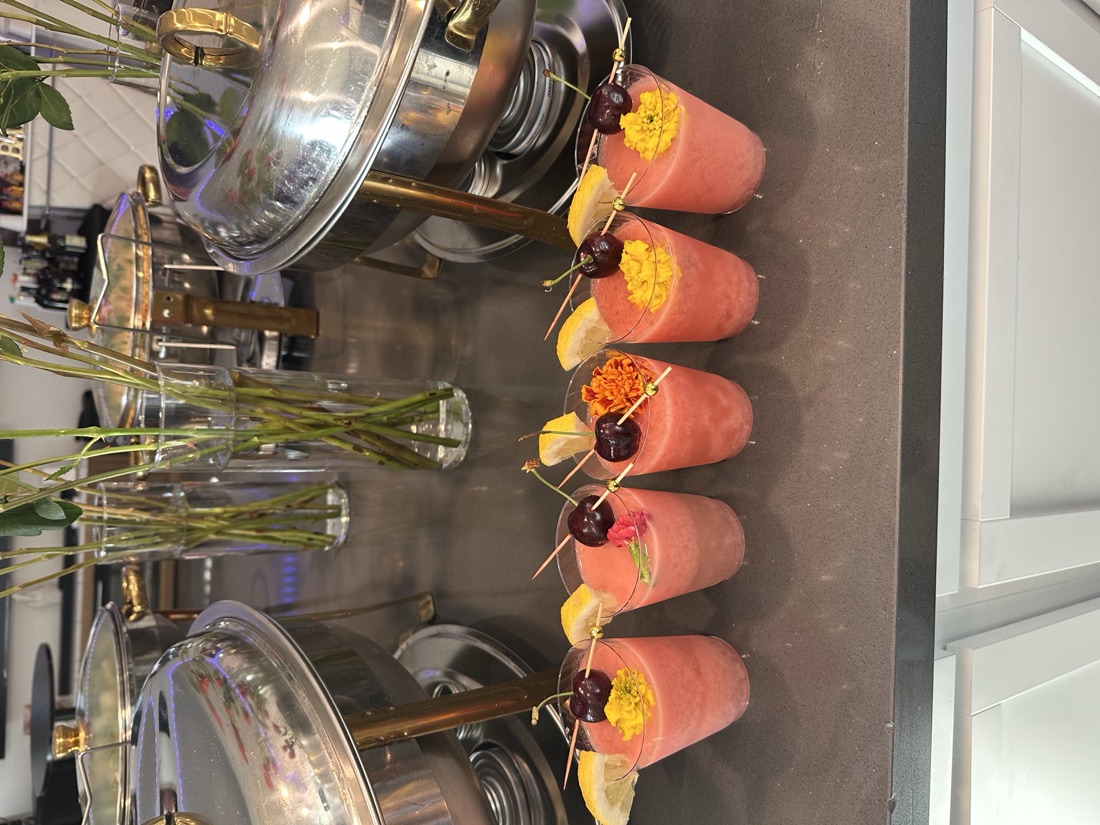 Five frozen watermelon-pink drinks garnished with cherries, lemon wedges, and marigold blooms lined up on a bar counter